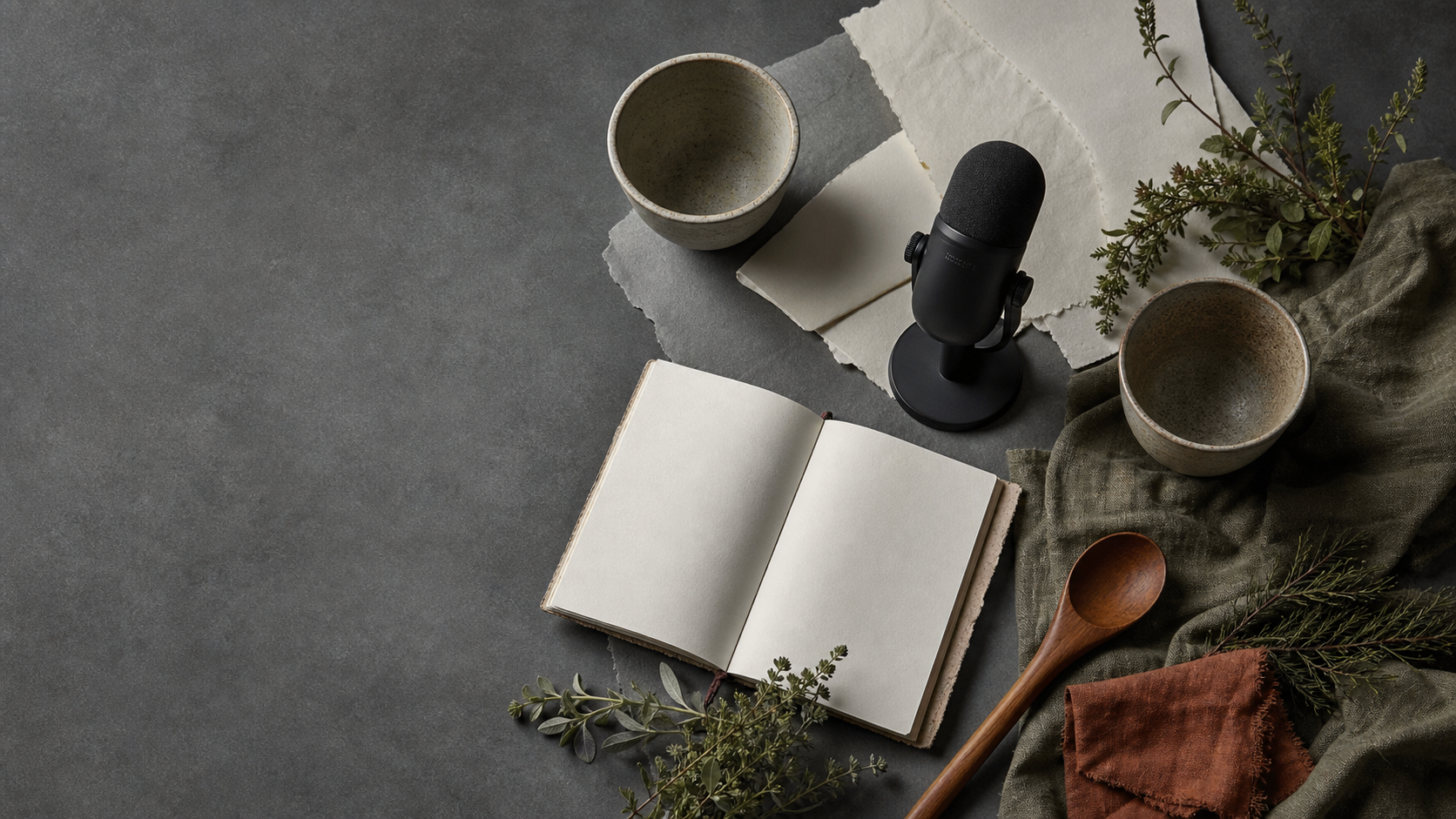 Overhead still life of a tabletop microphone, blank notebook, ceramic cups, herbs, cedar spoon, and rust linen arranged on a charcoal mineral paper table.